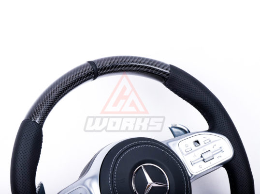 An upper section view of a 2019+ generation amg retrofit steering wheel, customized in gloss black carbon fiber, black perforated leather side grips, black nappa leather airbag and satin silver equipment.